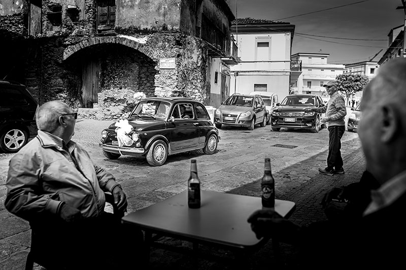 sposi in una 500 antica nera che passano davanti degli anziani al bar. Scatto di Mario Amelio Fotografo di matrimonio reportage.