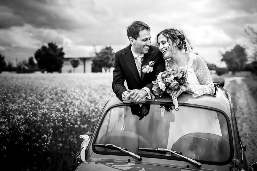 Sposi felici su un'auto d'epoca in un campo fiorito. Foto scatta da al fotografo matrimonio bergamo.
