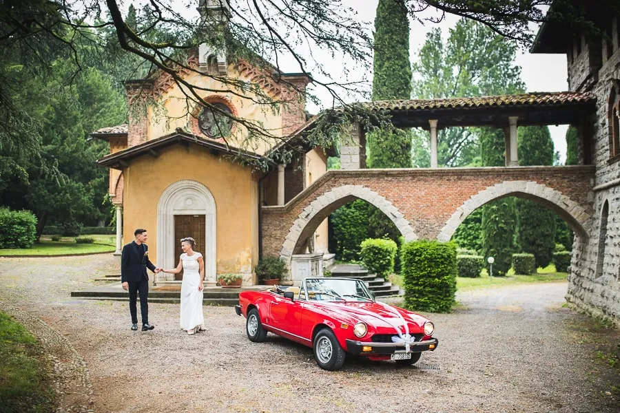 Sposi davanti alla chiesa di Villa Suardi a Bergamo, con un’auto d’epoca rossa a rendere l’atmosfera ancora più suggestiva. Scatto artistico di fotografo matrimonio bergamo.