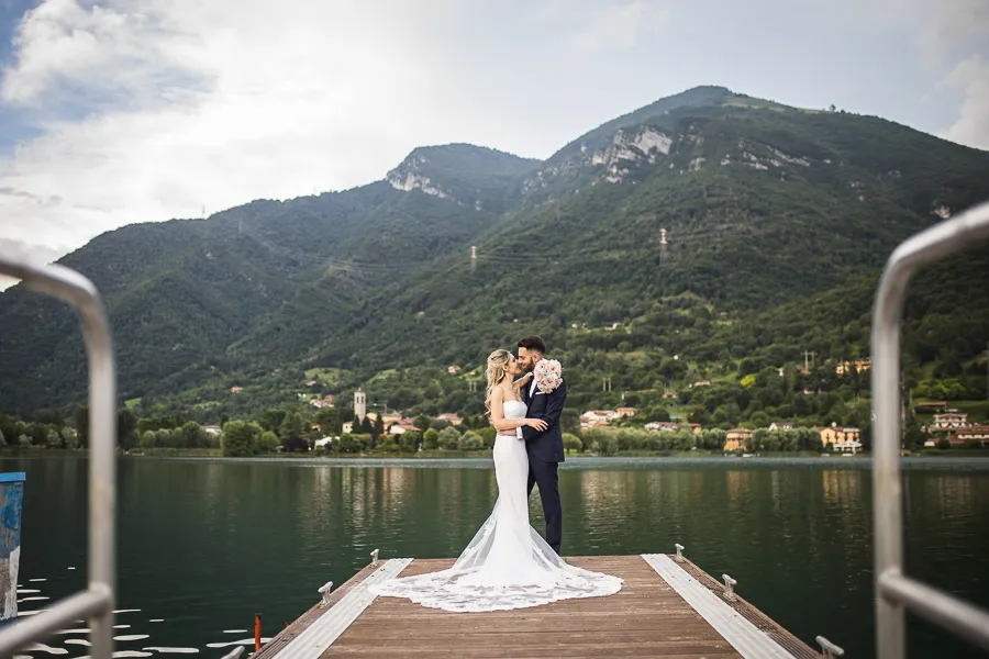 Gli sposi si abbracciano romanticamente sul molo con il lago e le montagne sullo sfondo a La Capannina al Lago, Bergamo
