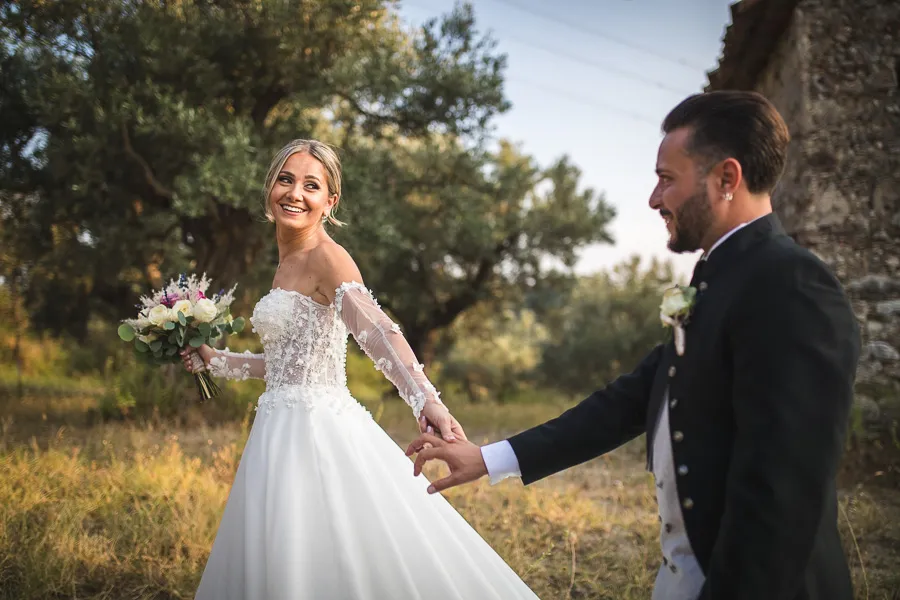 La sposa sorride mentre tiene per mano lo sposo in un uliveto illuminato dal sole
