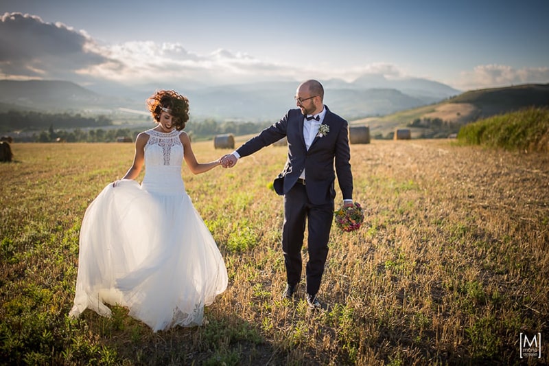 Gli sposi passeggiano mano nella mano in un campo al tramonto, tra balle di fieno e colline