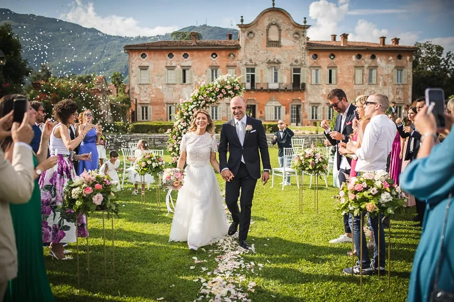 Sposi felici camminano mano nella mano dopo il rito civile all'aperto in una villa storica a Bergamo, un esempio di quanto il costo fotografo matrimonio Bergamo possa garantire ricordi indimenticabili.