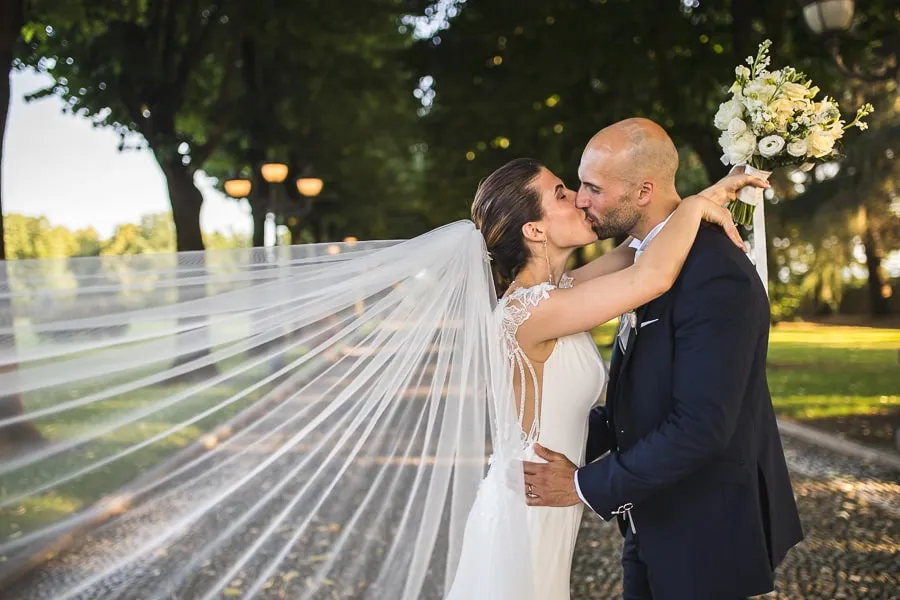 Bacio romantico degli sposi a Villa Valenca Fotografo matrimonio Brescia ritrae gli sposi mentre si scambiano un bacio romantico nel viale alberato di Villa Valenca, con il velo che si muove nel vento