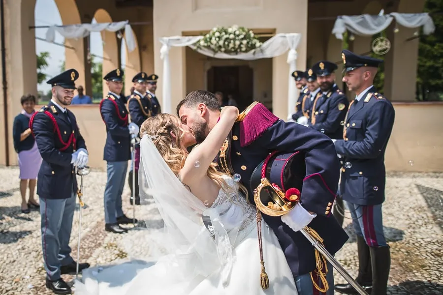 Bacio degli sposi con lo sposo in alta uniforme all’uscita della chiesa Fotografo matrimonio Brescia cattura il bacio emozionante degli sposi all’uscita della chiesa, con lo sposo in alta uniforme circondato dai colleghi