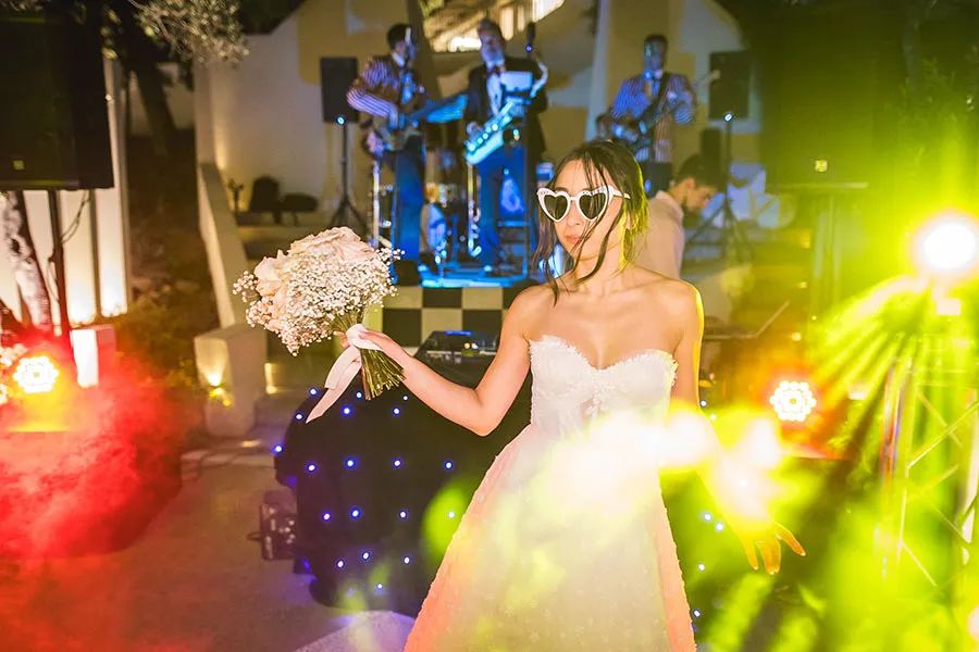Sposa con occhiali a cuore balla tra luci colorate con una band dal vivo sullo sfondo durante il ricevimento di nozze, evidenziando l'importanza della scelta del fotografo di matrimonio a Catanzaro.