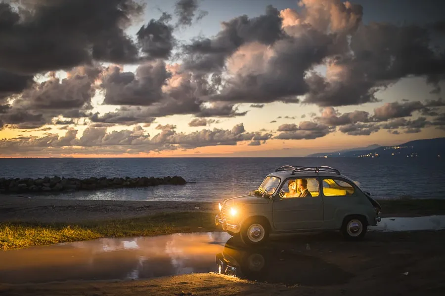 Sposi in auto vintage illuminati dai fari con sfondo marino, timing perfetto frutto della collaborazione fotografo wedding planner