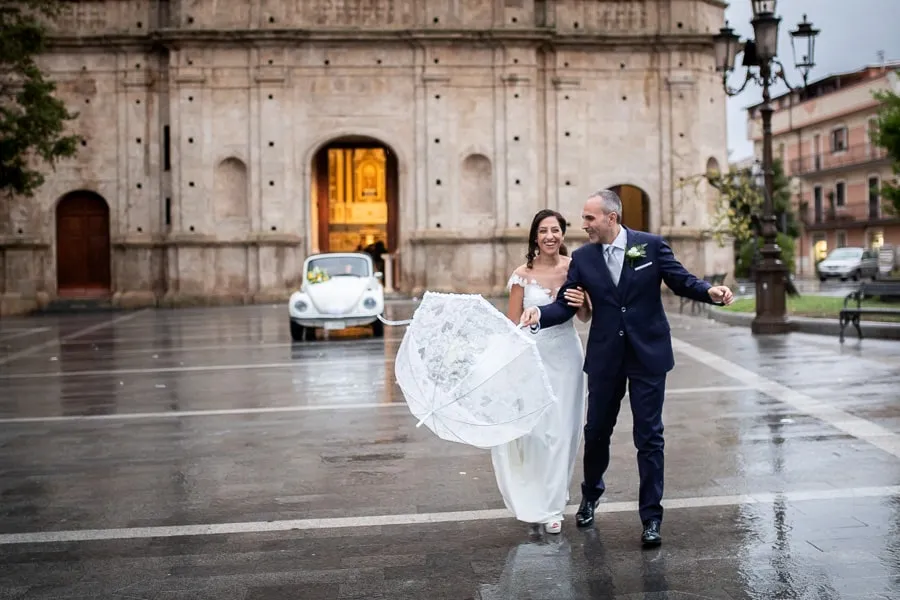 Sposi sorridenti camminano sotto la pioggia con l’ombrello davanti alla chiesa, dimostrazione di perfetta gestione imprevisti matrimonio