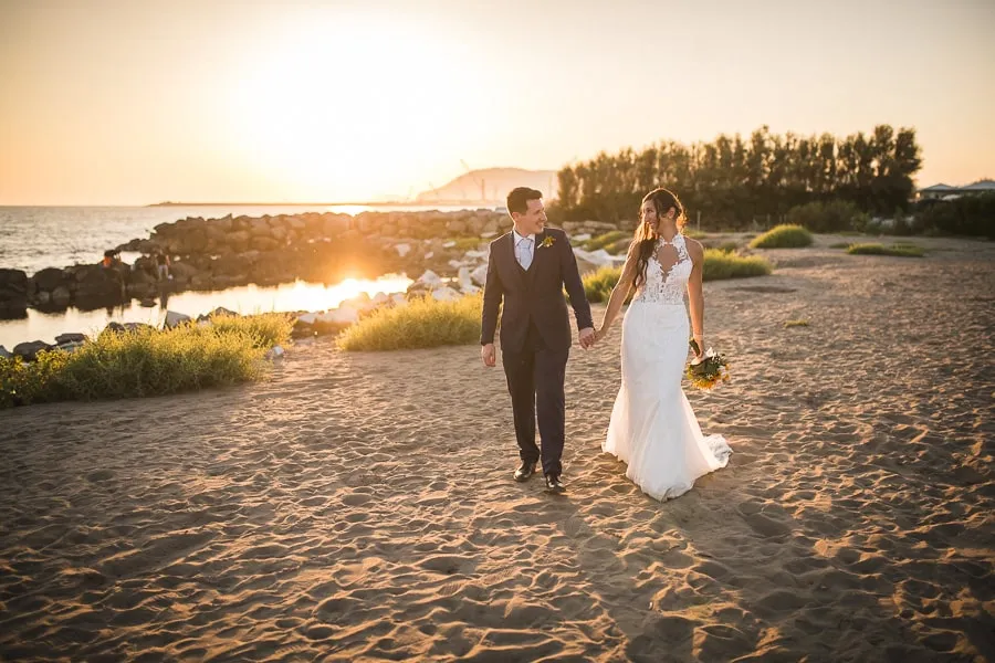 Sposi che camminano mano nella mano sulla spiaggia al tramonto, ideale per la selezione foto matrimonio